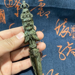 May include: A bronze-colored, ornate dagger with a detailed handle featuring a carved face and figures. The blade has a pointed tip and etched designs. The dagger is held against a blue patterned background.