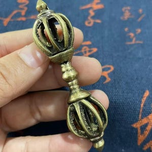May include: A bronze-colored vajra, a ritual object with a symmetrical design. The vajra features a central handle with ornate detailing and two symmetrical, multi-pronged ends. The object is held in a hand against a blue background.