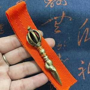 May include: A bronze-colored Vajra, a ritual object, rests on an orange fabric. The Vajra features a detailed design with a central sphere and pointed ends. The orange fabric is set against a blue background with script.