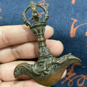 May include: A bronze-colored, ornate ritual object with a detailed handle and a curved, blade-like end. The object has intricate carvings and a weathered appearance, suggesting age and use. The design features a loop at the top and a flared base.