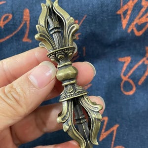 May include: A bronze-colored vajra, a ritual object, is held in a hand. The vajra features ornate, symmetrical designs with a central column and flared ends. The object has a weathered appearance, suggesting age or a vintage style.