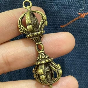 May include: A detailed brass pendant with an ornate design. The pendant features a crown-like top, a central section with intricate patterns, and a bell-shaped bottom. The overall design is symmetrical and detailed, with a vintage aesthetic.