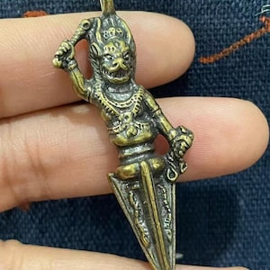 May include: A bronze-colored pendant featuring a detailed figure holding a weapon, with a triangular blade at the base. The figure has an ornate headdress and intricate detailing on the body. The pendant is designed with a loop for hanging.