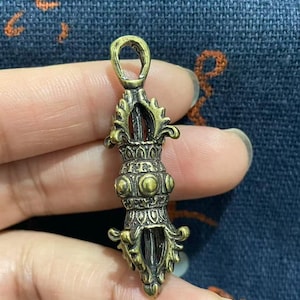 May include: A detailed, antique-style brass pendant with intricate carvings. The pendant features a loop at the top for hanging and a symmetrical design with ornate details. The color is a warm, aged brass.