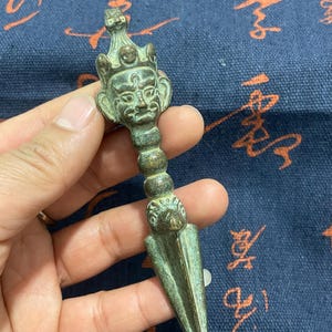 May include: A bronze-coloured, ornate dagger with a detailed face at the handle's top. The dagger has a pointed blade and a decorative handle. The object is held in a hand against a blue background with Asian script.