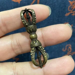 May include: A bronze-colored vajra, a ritual object, held in a hand. The vajra features ornate detailing, with a central rod and decorative ends. The object is likely used in Buddhist or Hindu rituals.