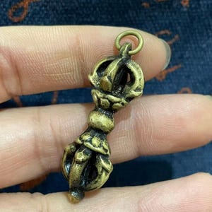 Pode incluir: Um pequeno pingente vajra de latão em estilo antigo com trabalho de metal simétrico e intrincado, uma empunhadura central e extremidades pontiagudas. O objeto inclui um anel circular no topo para fixação e apresenta um acabamento metálico dourado e desgastado.