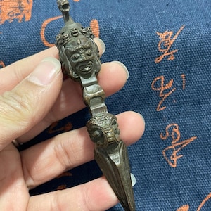May include: Bronze-colored ritual dagger with intricate carvings. The handle features a detailed face with a crown, and the blade tapers to a sharp point. The dagger is held against a blue patterned background.