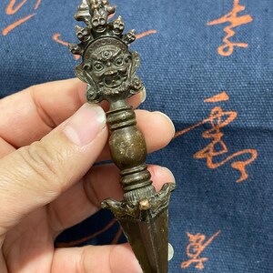 May include: A bronze-colored, ornate dagger with a detailed handle featuring a face with multiple eyes and a crown. The dagger has a triangular blade and is held in a hand against a blue patterned background.