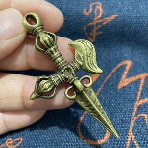 May include: A detailed bronze-colored Vajra pendant. The pendant features ornate detailing, including a central cross-like design, curved handles, and a pointed blade. The top has a loop for hanging. The overall design is symmetrical and intricate.