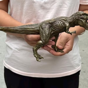 May include: A bronze-colored Tyrannosaurus rex dinosaur figurine with a textured, scaly surface. The dinosaur is depicted in a standing position with its mouth open, showing teeth, and its long tail extended horizontally to the side.