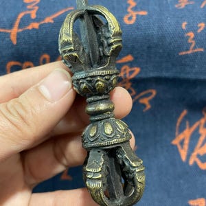 May include: A detailed bronze vajra, a ritual object with ornate details. The vajra features a central sphere, two lotus-shaped bases, and intricate designs. The object is held against a blue background with Asian script.