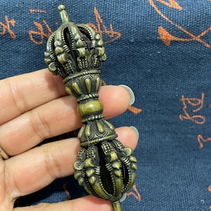 May include: A detailed bronze-colored vajra, a ritual object, held in a hand. The vajra features intricate crown-like designs at both ends and a central grip. The object is approximately 6 inches in length.