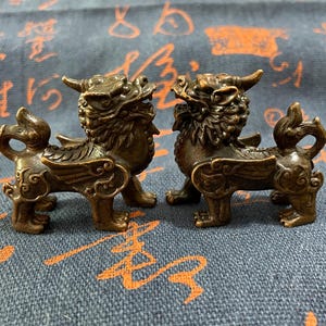 May include: Two bronze-colored mythical creatures, possibly Foo Dogs or similar guardians, are displayed. Each figurine has a detailed design, including wings, horns, and a lion-like mane. They are positioned side-by-side on a patterned blue surface.
