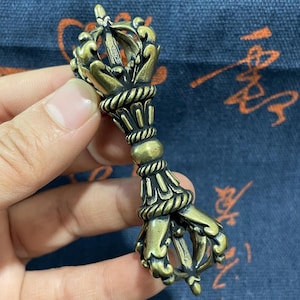May include: A detailed bronze-coloured Vajra, a ritual object, featuring intricate floral and rope-like designs. The ends are shaped like stylised hands, and the centre has a decorative band. This item is likely used in religious or spiritual practices.