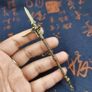 May include: A bronze-colored miniature spear with intricate detailing. The spearhead features a blade and a hook, while the shaft is adorned with ornate patterns. The spear is held in a hand, showcasing its size and craftsmanship.