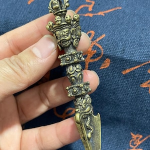 May include: A metallic bronze-colored phurba ritual dagger featuring an intricate handle with a wrathful deity face at the top, decorative bands, and a sharp, pointed blade at the bottom. The object displays detailed relief carvings throughout.