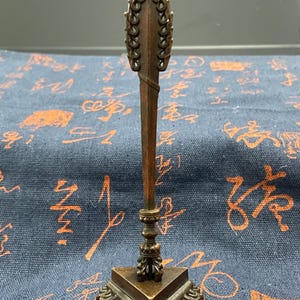 May include: A bronze-colored, ornate decorative object with a spear-like top. The spearhead is detailed with a series of connected loops. The base is square with a decorative edge. The object stands on a blue patterned surface.