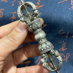 May include: A silver Vajra, a ritual object with ornate floral and geometric designs. The ends feature a trident shape, and the handle has a bulbous shape with lotus flower details. The object is held in a hand.
