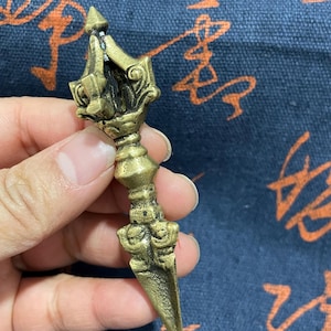 May include: An antique bronze dagger with an ornate handle and a sharp, pointed blade. The dagger is held in a hand against a blue background with orange patterns. The design features detailed carvings and a decorative top.