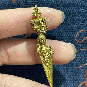 May include: A gold-colored, ornate dagger with a detailed handle featuring multiple faces and a crown. The blade is triangular and tapers to a point. The dagger is held in a hand against a dark blue background.
