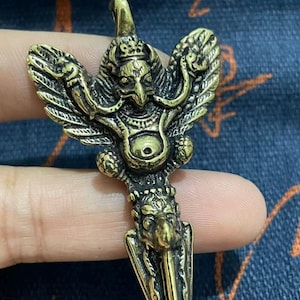 May include: A detailed brass pendant featuring a bird-like deity with outstretched wings and a pointed blade at the bottom. The pendant has intricate carvings and a loop at the top for hanging. The color is a warm, antique gold.