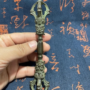 May include: A bronze-colored, ornate ritual object with a detailed design. The object features a central handle with two faces and intricate patterns. The top has a crown-like structure. The overall design suggests a religious or ceremonial purpose.