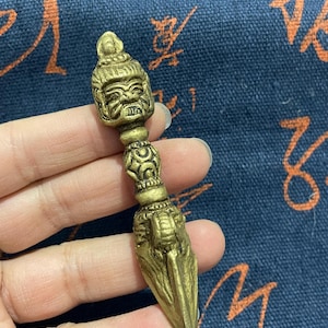 May include: A detailed brass ritual dagger, featuring a carved deity head at the handle and a stylized blade. The dagger is held against a blue patterned fabric background, showcasing its intricate design and golden colour.