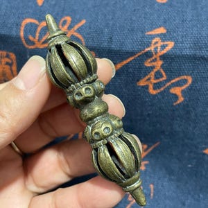 May include: A bronze-colored, ornate metal object with a detailed, symmetrical design. The object has a central column with bulbous sections and a pointed top and bottom. The intricate details suggest a religious or ceremonial item.