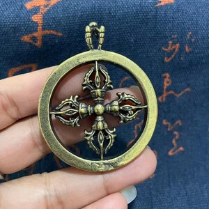 May include: A circular brass-toned metal pendant featuring a central four-pronged vajra symbol. The design is enclosed within a smooth, round outer frame with a small integrated bail at the top for attachment, showcasing a weathered, antique finish.