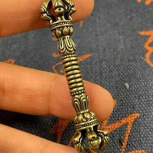 May include: A detailed, antique-style brass vajra, a ritual object with ornate detailing. The vajra features a central ribbed section and decorative ends with intricate patterns. The overall design is symmetrical and the brass has a warm, aged appearance.