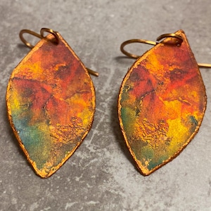 May include: A pair of leaf-shaped earrings featuring a textured surface with a gradient of blended colors including deep red, bright orange, yellow, and hints of teal. Each earring is suspended from a simple copper-toned metal hook wire.