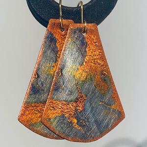 May include: A pair of trapezoidal drop earrings featuring a textured, metallic finish with blended hues of copper, blue, and gold. The earrings are suspended from thin, curved gold-toned metal hooks, showcasing a brushed surface with visible color variations.