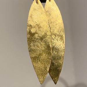 May include: A pair of elongated, leaf-shaped gold-toned earrings with a textured, hammered surface. Each earring features a pointed tip and is suspended from a thin gold-toned wire hook, showcasing a metallic finish with visible surface variations.