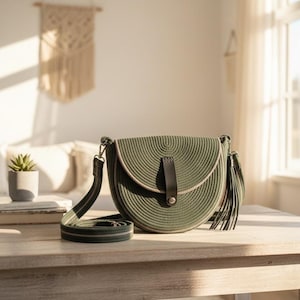 Può includere: Borsa a tracolla verde oliva con un design intrecciato circolare. La borsa presenta un cinturino e un nappa in pelle nera e una tracolla regolabile abbinata. La borsa è su un tavolo di legno.