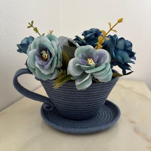 Può includere: Una tazza e un piattino decorativi in corda blu navy, riempiti di fiori artificiali blu, turchese e viola, con dettagli gialli. La tazza ha un manico e poggia su un piattino abbinato.