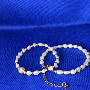 May include: Two gold and white pearl bracelets with a small gold charm. The bracelets are on a blue background.