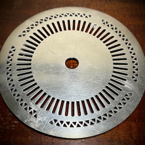 May include: A circular metal disc with a series of evenly spaced slots and triangular cutouts around the edges. The disc has a central hole and a brushed metal finish. The object is likely a component for machinery or a decorative element.