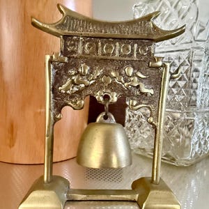 May include: A brass decorative bell suspended from a frame shaped like a traditional East Asian gate. The frame features a textured surface with embossed figures and a smooth, polished bell hanging from a central loop on a stable base.