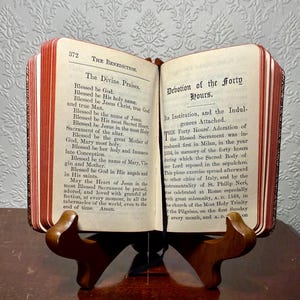 May include: An open antique book with red-edged pages, displayed on a wooden stand. The book's pages feature religious text, including "The Divine Praises" and "Devotion of the Forty Hours." The book is open to reveal the text.