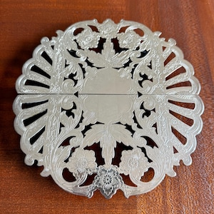 May include: A silver-colored, ornate trivet with a floral and leaf design. The trivet is round with a scalloped edge and a hinged, rectangular center. The design includes detailed floral patterns and openwork sections.