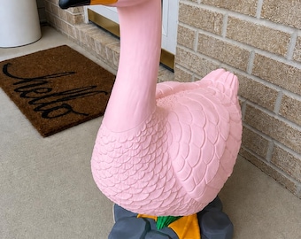 May include: A large, standing pink goose figurine with a bright orange beak and a black tip. The goose has textured, sculpted feathers and is positioned on a base of gray, faceted rocks. The eyes are small, round, and black in color.