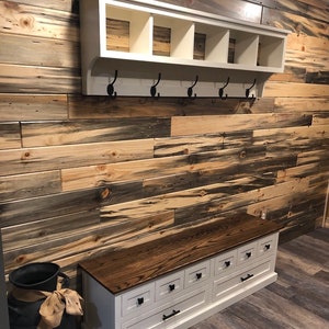 May include: White wooden bench with six drawers and a dark wood top. The bench is against a wall with blue and brown wood paneling. A white shelf with hooks is mounted above the bench.