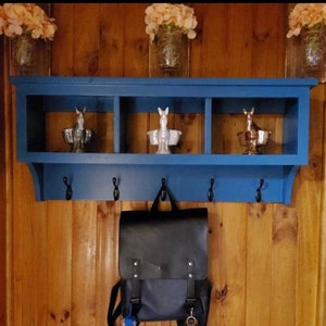 May include: A blue wooden shelf with three cubbies and five hooks. The shelf has three decorative ceramic figurines on the top. A black leather backpack is hanging on one of the hooks.