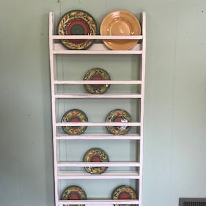 May include: White wooden plate rack with seven shelves, holding colourful plates with floral designs. The rack is against a light green wall.