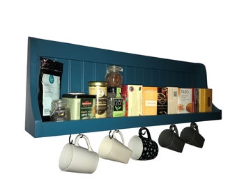 May include: A teal blue wall-mounted shelf with a wooden top and metal hooks underneath. The shelf is filled with various tea boxes, jars, and cans. There are five mugs hanging from the hooks.
