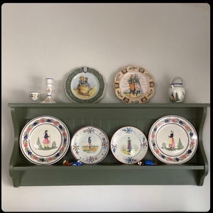 May include: A green wooden shelf with five decorative plates. The plates are white with blue and red designs. The plates are arranged in a row on the shelf. There is a small white ceramic pitcher on the shelf.