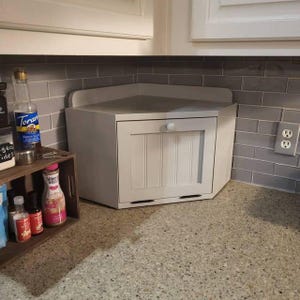 May include: A light gray corner bread box with a vertical paneled door and a small round knob, positioned on a speckled countertop next to a wooden shelf containing various bottles of syrup and a bottle of Torani brown sugar cinnamon syrup.