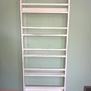 May include: White wooden shelf unit with seven shelves. The shelf unit is standing against a light green wall.