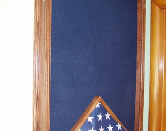 May include: A wooden flag display case with a dark blue background and a folded American flag. The case has a rectangular shape with a hinged front and a brown wooden frame. The flag is displayed in a triangular shape, with white stars on a blue field.